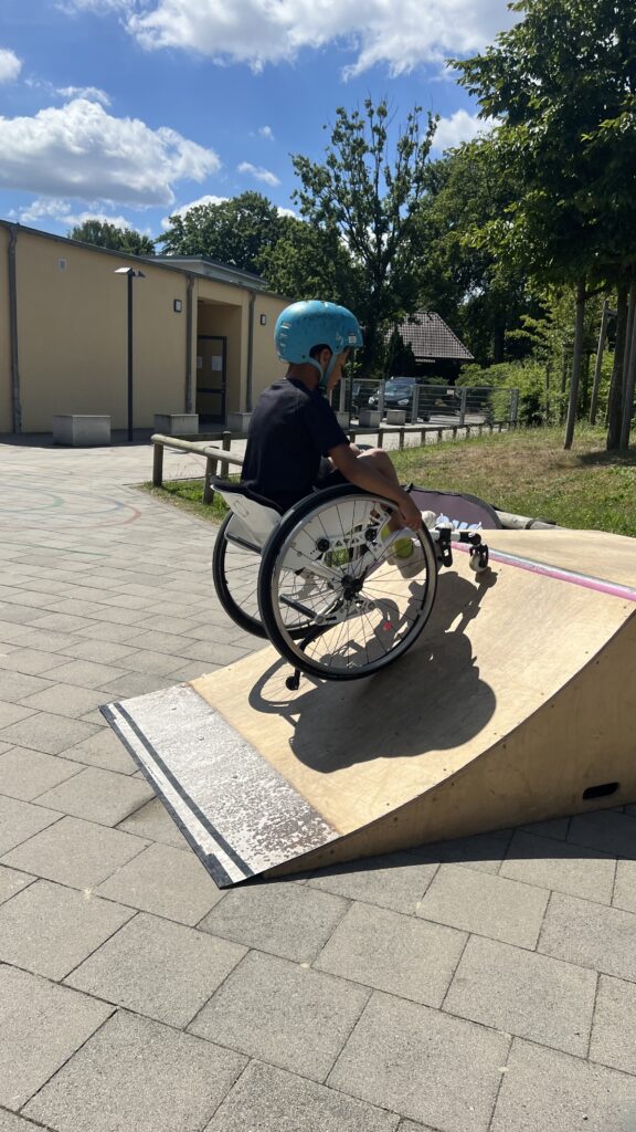 Ein Junge im Rollstuhl mit blauem Helm fährt konzentriert eine kleine Rampe hoch. Die Szene spielt sich draußen auf einem Schulhof bei sonnigem Wetter ab. Im Hintergrund steht ein flaches Schulgebäude.