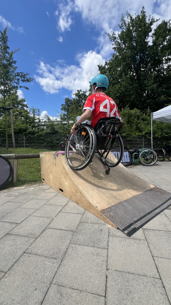 Ein Jugendlicher mit blauem Helm und rotem Trikot („FC Bayern“) fährt einen mobilen Skate-Ramp hoch. Er sitzt im Rollstuhl und schaut nach vorne, während der Himmel mit Wolken und Bäumen im Hintergrund zu sehen ist.