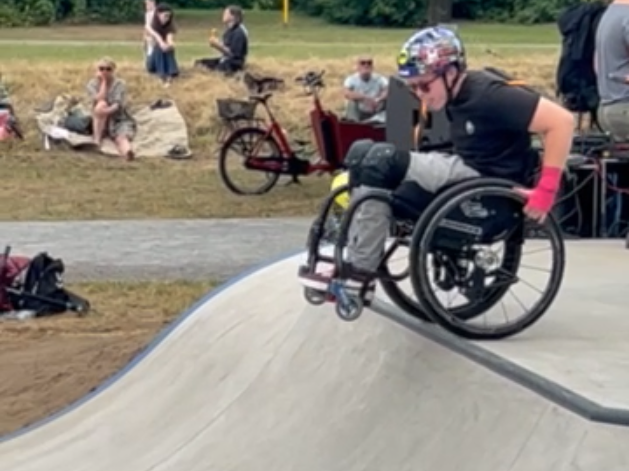 Eine Person im Rollstuhl fährt auf einem Beton-Skatepark über eine abgerundete Rampe. Sie trägt Helm, Schoner und pinke Handschuhe. Im Hintergrund sitzen und stehen Zuschauer*innen auf einer Wiese.