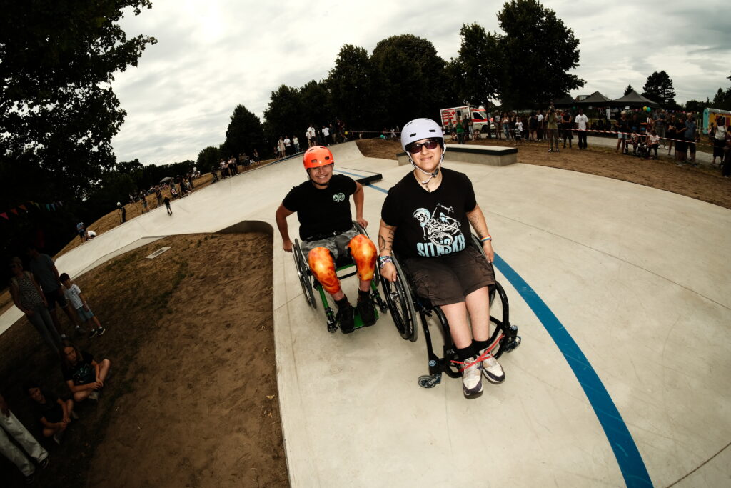 Zwei Personen im Rollstuhl fahren nebeneinander durch den Skatepark. Beide tragen Helme, lächeln in die Kamera und tragen SIT’N’SKATE-Shirts. Hinter ihnen stehen Menschen am Rand und beobachten das Geschehen.