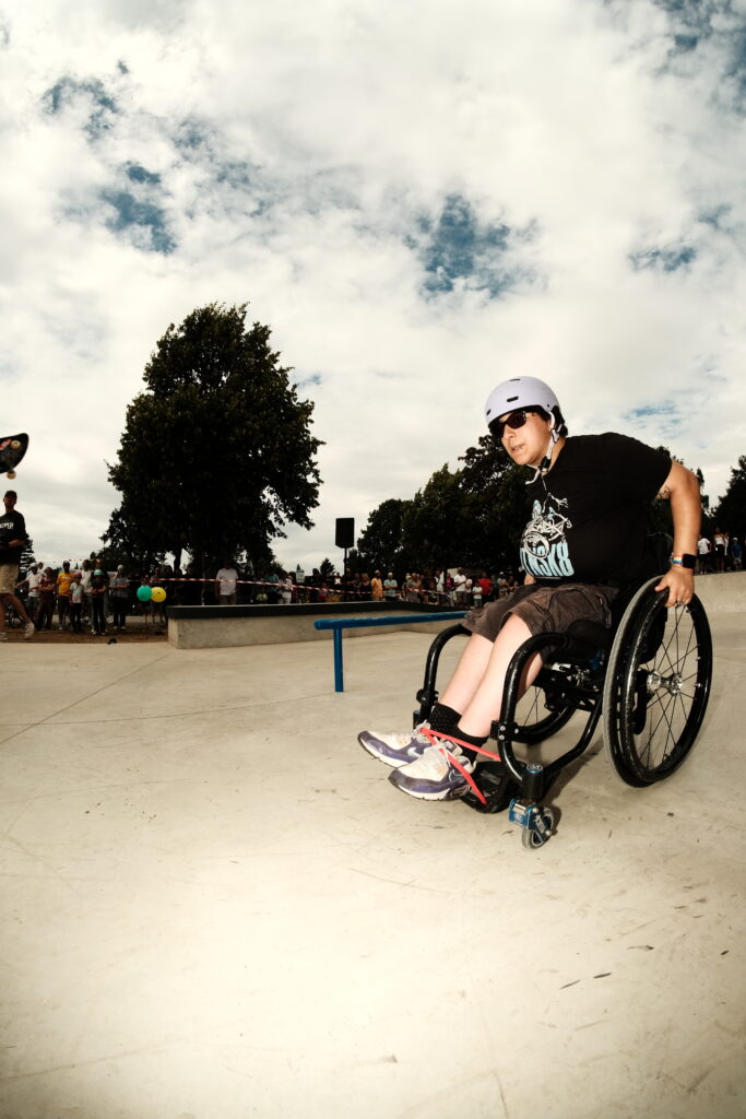 Eine Person im Rollstuhl trägt Helm und Sonnenbrille und steht im Skatepark vor einem blauen Rail. Im Hintergrund sind viele Menschen, Bäume und ein wolkiger Himmel zu sehen.