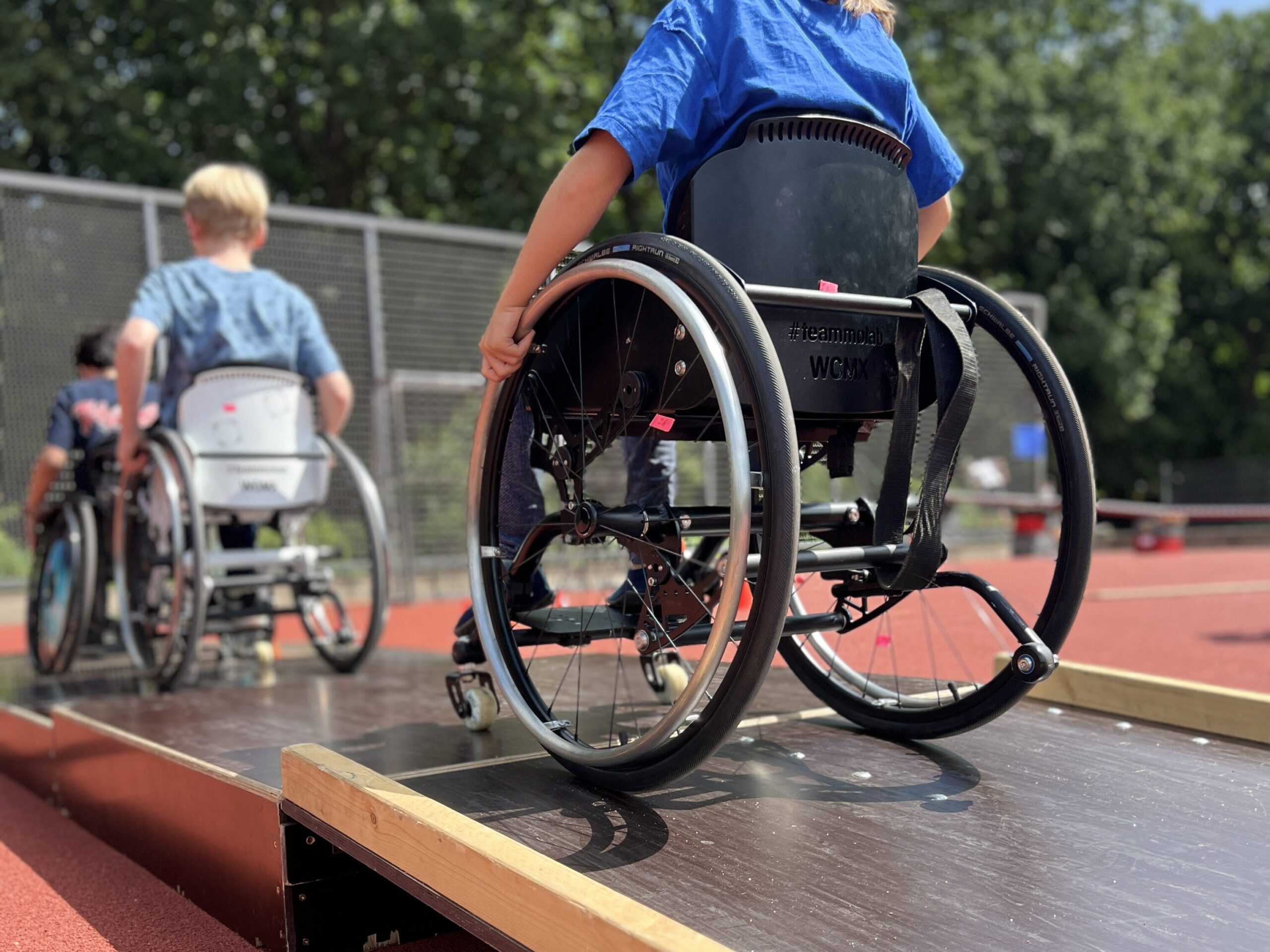Kinder im Rollstuhl fahren über eine mobile Rampe auf einem roten Sportplatz. Im Vordergrund ist ein Kind mit blauem T-Shirt im Rollstuhl zu sehen, das gerade die Rampe hinunterfährt. Weitere Kinder im Rollstuhl sind auf dem Weg nach oben. Im Hintergrund sieht man einen Zaun und grüne Bäume.