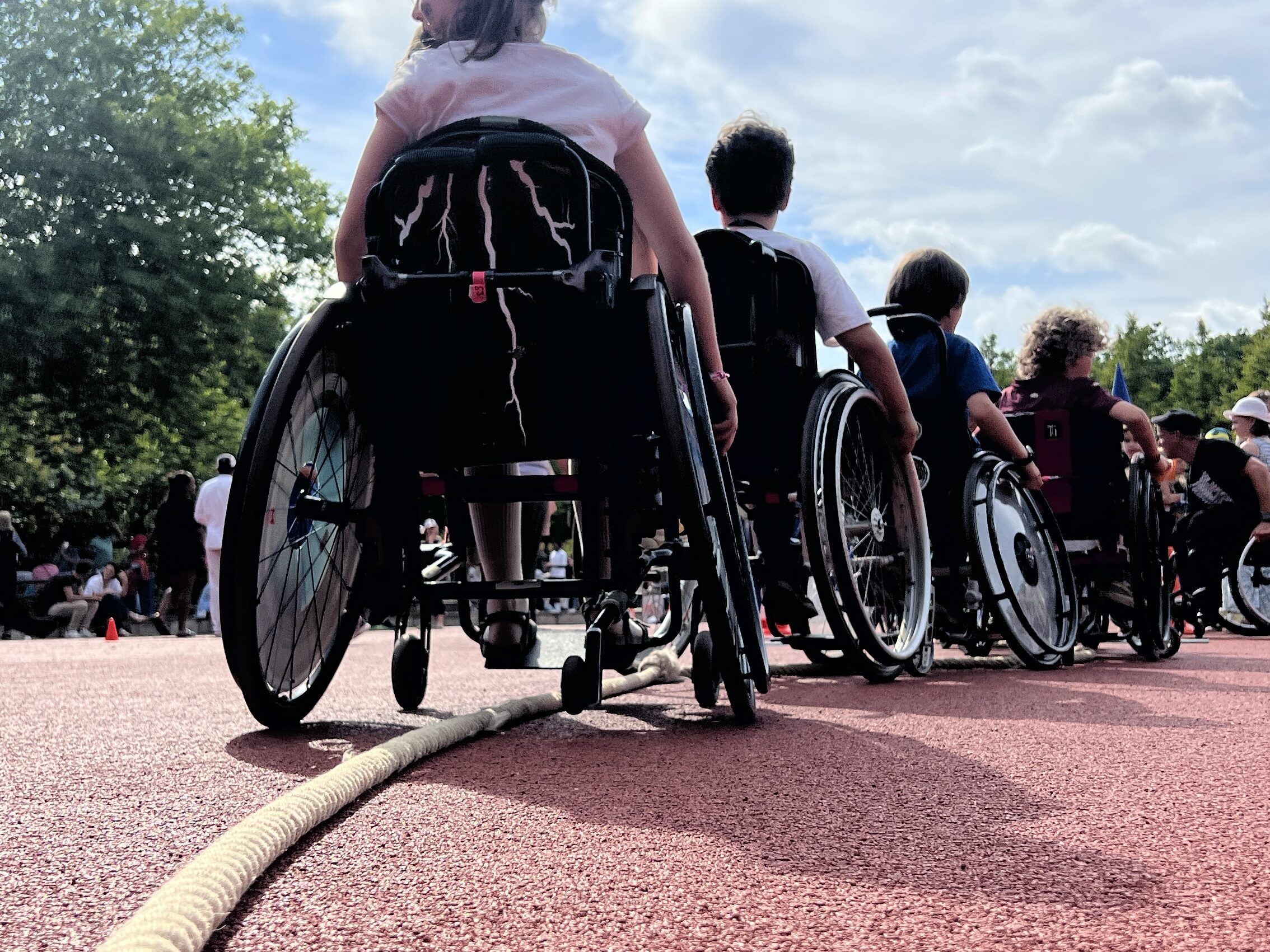 Eine Gruppe von Kindern im Rollstuhl rollt gemeinsam über ein dickes Tau. Die Perspektive zeigt die Kinder von hinten, während sie sich auf dem roten Sportplatz mit ihren Rollstühlen in eine Richtung bewegen. Um sie herum stehen weitere Personen. Es ist