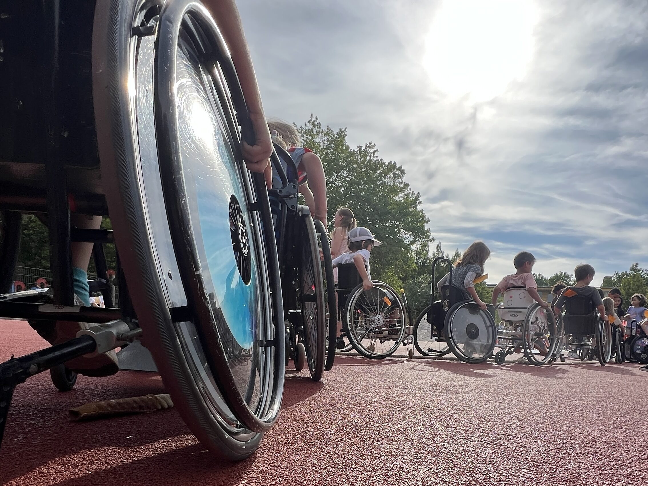 Nahaufnahme eines Rollstuhlrads mit silbernem Speichenschutz. Mehrere Kinder im Rollstuhl sind auf einem roten Sportplatz zu sehen, die Sonne scheint durch einen bewölkten Himmel. Im Hintergrund sitzen und fahren weitere Kinder im Rollstuhl. Die Perspektive ist bodennah und betont die Bewegung und Dynamik.
