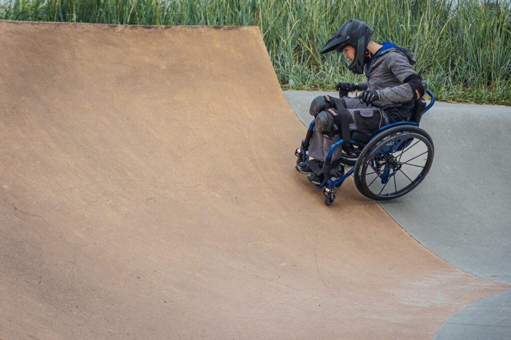 Eine jugendliche Person im Rollstuhl mit Helm, Protektoren und Handschuhen fährt eine steile Betonrampe im Skatepark hinunter. Die Szene ist ruhig, im Hintergrund sind hohe Gräser zu sehen.
