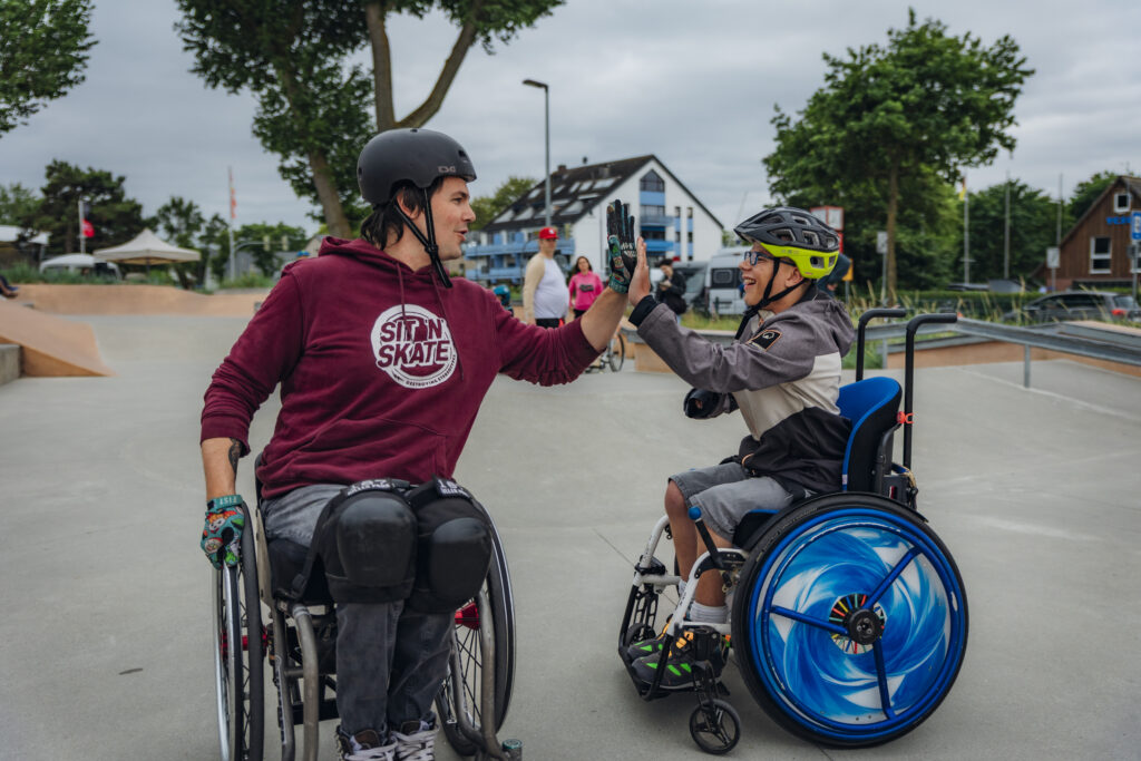 Zwei Rollstuhlfahrende, ein erwachsener Mann im SIT’N’SKATE-Hoodie und ein Kind mit Helm und bunter Radkappe, geben sich ein freudiges High Five im Skatepark. Beide lächeln – im Hintergrund sind Rampen, Bäume und weitere Menschen zu sehen.