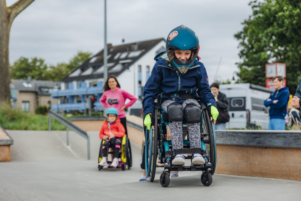 Ein Kind im Rollstuhl fährt mit Helm und Protektoren konzentriert eine Skateparkrampe hinunter. Dahinter ein weiteres Kind im Rollstuhl sowie eine Frau in pinkem Hoodie, die beide beobachten. Weitere Personen schauen von der Seite zu.