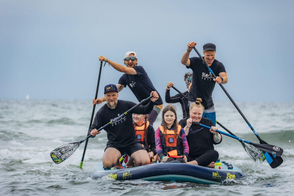 Acht Personen auf einem großen Stand-Up-Paddle-Board auf dem offenen Meer. Vorne sitzen drei Kinder mit Schwimmwesten, hinten stehen fünf Erwachsene, die mit Paddeln das Board manövrieren. Das Wasser ist wellig, der Himmel bedeckt – alle haben sichtlich Spaß.
