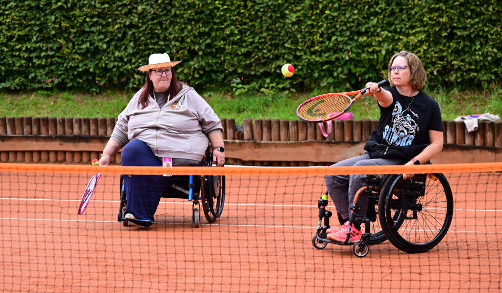 Zwei Frauen im Rollstuhl spielen gemeinsam Tennis. Eine trägt einen Sonnenhut, die andere ein SIT’N’SKATE-T-Shirt und schlägt den Ball mit dem Schläger über das Netz.