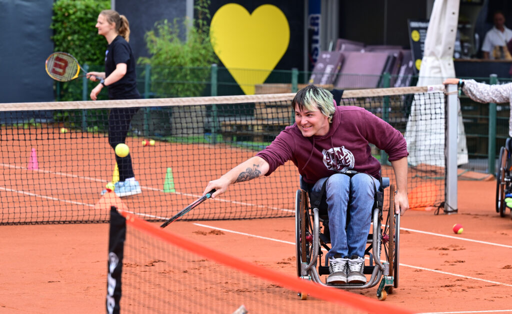 David sitzt im Rollstuhl auf dem Tennisplatz, streckt den Arm weit nach vorne und trifft den Ball mit dem Schläger. Er trägt einen bordeauxfarbenen SIT’N’SKATE-Hoodie und lächelt konzentriert.