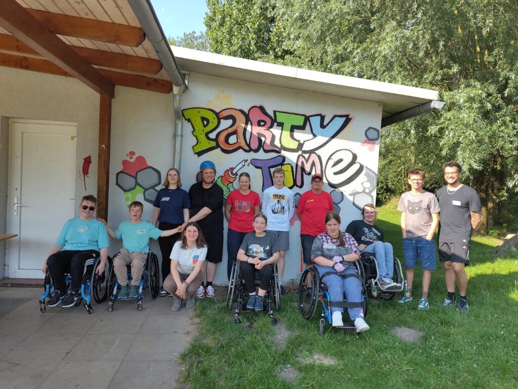 Gruppenfoto vor einer bemalten Wand mit dem bunten Schriftzug „Party Time“. Zu sehen sind 15 Personen: Kinder, Jugendliche und Erwachsene – einige im Rollstuhl, andere stehend. Sie stehen oder sitzen fröhlich nebeneinander auf einer Wiese vor einem kleinen Gebäude mit Holzvordach. Die Stimmung wirkt entspannt und freundlich, die Sonne scheint. Das Bild entstand beim inklusiven Ferienangebot im Funpark Bremen.