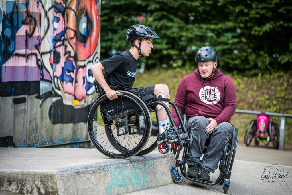 Zwei Rollstuhlfahrende stehen an einer Kante im Skatepark. Links ein junger Teilnehmer mit schwarzem Helm, schwarzem T-Shirt und Orthesen an den Beinen, der seinen Rollstuhl an der Kante positioniert. Rechts ein Erwachsener mit schwarzem Helm und rotem SIT’N’SKATE-Hoodie, der Tipps gibt.