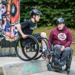 Zwei Rollstuhlfahrende stehen an einer Kante im Skatepark. Links ein junger Teilnehmer mit schwarzem Helm, schwarzem T-Shirt und Orthesen an den Beinen, der seinen Rollstuhl an der Kante positioniert. Rechts ein Erwachsener mit schwarzem Helm und rotem SIT’N’SKATE-Hoodie, der Tipps gibt.