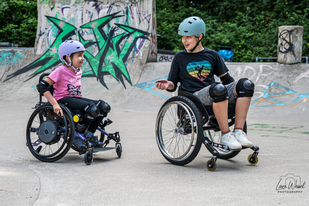 Zwei Kinder im Rollstuhl unterhalten sich lachend im Skatepark. Das Mädchen links trägt einen lilafarbenen Helm, pinkes T-Shirt und schwarze Schoner. Der Junge rechts trägt einen hellblauen Helm, ein schwarzes T-Shirt mit buntem Herzmotiv und Knieschoner.