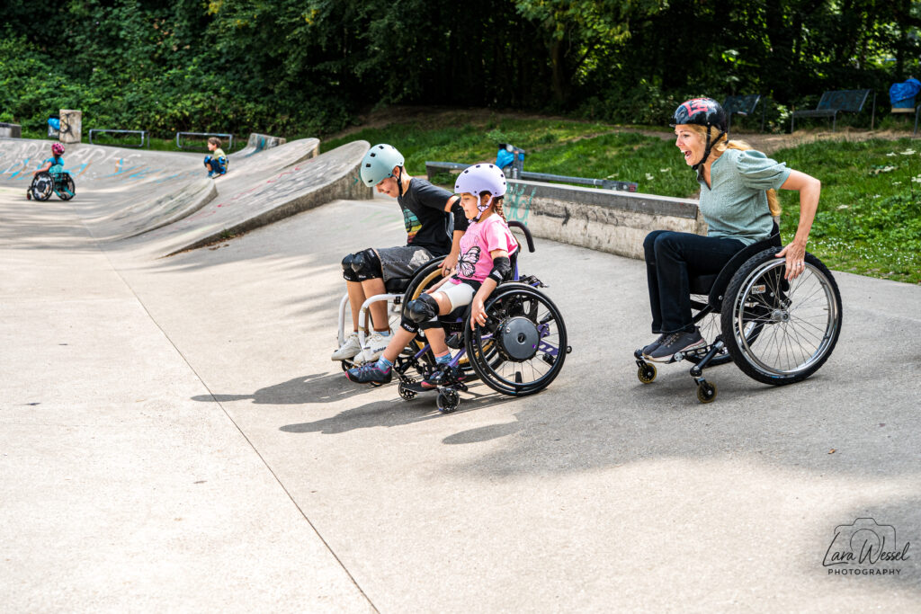 Drei Rollstuhlfahrende fahren nebeneinander über die Fläche des Skateparks. Links ein Junge mit hellblauem Helm, in der Mitte ein Mädchen mit lilafarbenem Helm, rechts eine Frau mit rotem Helm und grünem Oberteil, die lachend anschiebt.