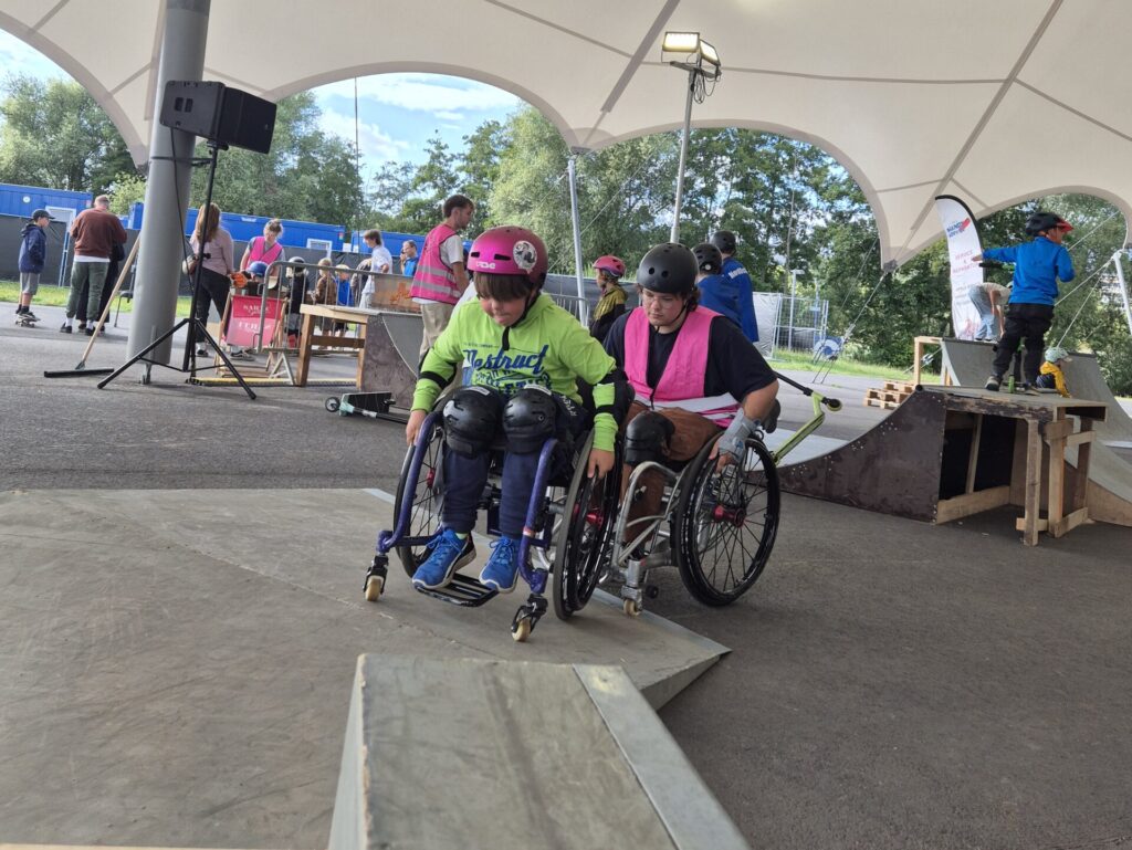 Zwei Menschen im Rollstuhl mit Helmen und Schutzausrüstung fahren hintereinander eine Rampe im Skatepark rauf. Im Hintergrund sind weitere Menschen zu sehen, die zuschauen oder selbst aktiv sind.
