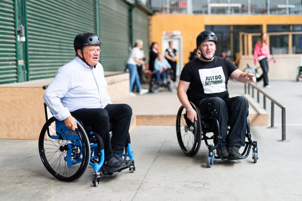 Minister Philippi im Gespräch mit einem erfahrenen Rollstuhlfahrer in der Skatehalle. Beide sitzen sich im Rollstuhl gegenüber und tragen Helme. Im Hintergrund sind Rampen und Elemente der Halle sichtbar.