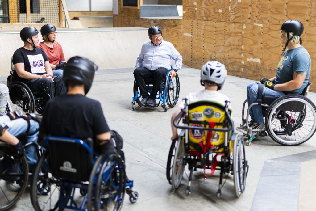 Andreas Philippi sitzt im Rollstuhl in einer Gruppe von Rollstuhlfahrenden mitten in der Skatehalle. Die Teilnehmenden tragen Helme und bilden einen Kreis. Es wirkt wie eine Gesprächs- oder Einführungsrunde.