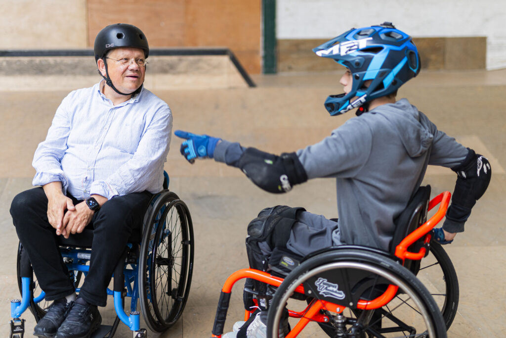 Ein junger Rollstuhlfahrer mit Helm und Schutzausrüstung zeigt lächelnd auf Minister Philippi, der ihm im Rollstuhl gegenübersitzt. Die Szene wirkt locker und kommunikativ.