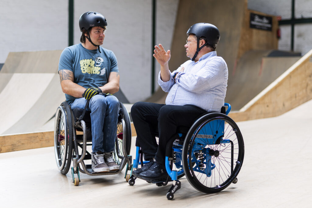 Minister Philippi im Rollstuhl im Gespräch mit David Lebuser. Beide tragen Helme. Im Hintergrund sind weitere Personen und Elemente der Skatehalle zu erkennen.