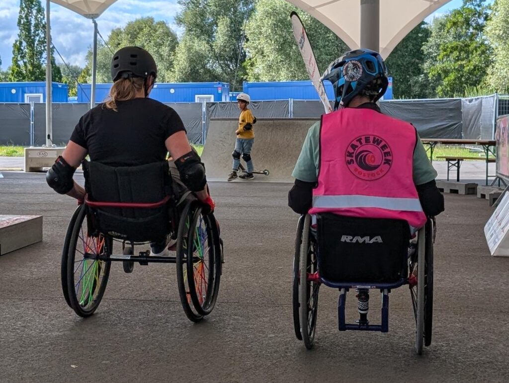 Zwei Menschen im Rollstuhl mit Helmen rollen nebeneinander durch den überdachten Skatepark. Einer trägt ein pinkes Skateweek-Westen-Shirt, im Hintergrund ist ein Kind mit Skateboard zu sehen.