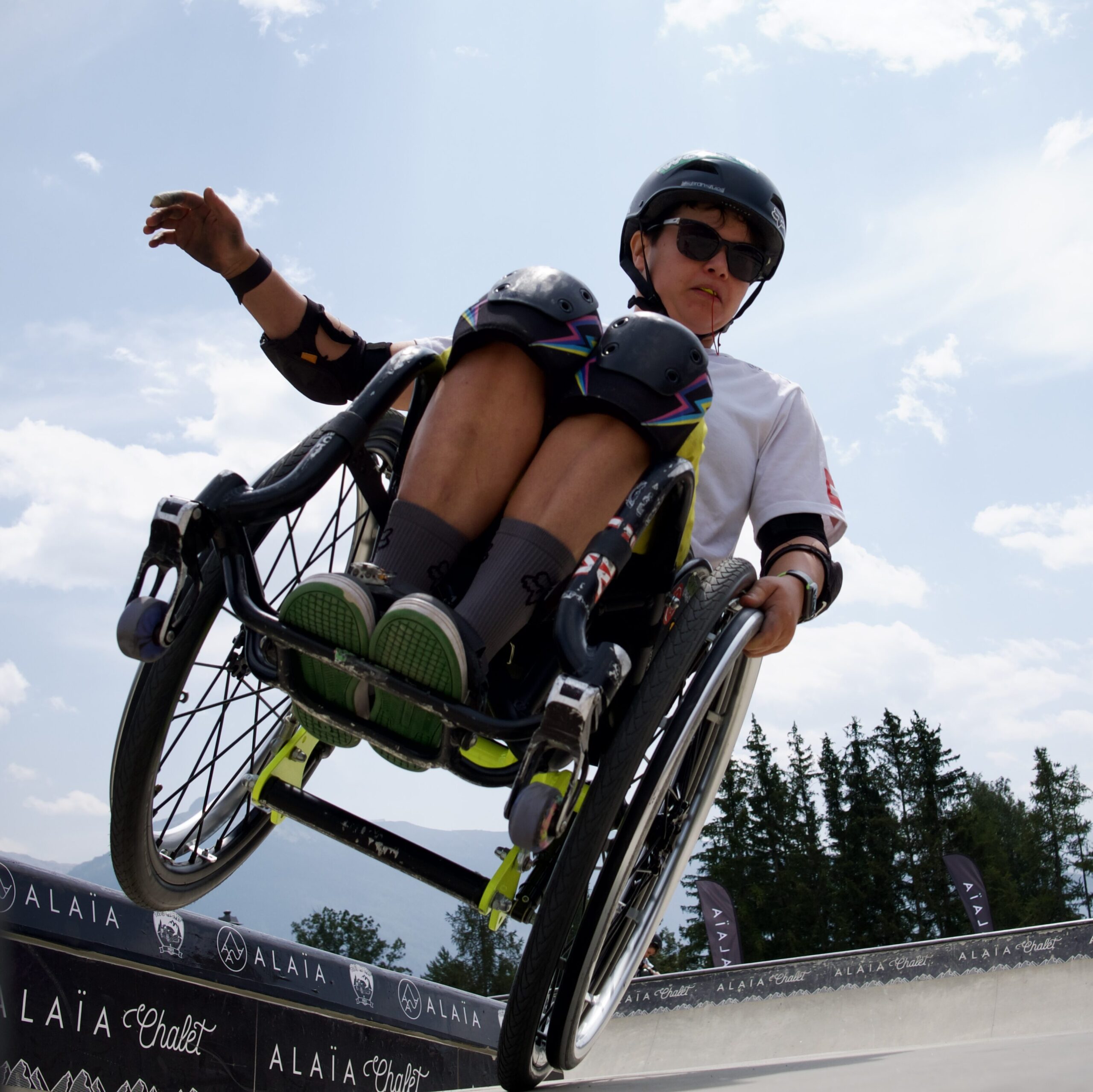 Eine Rollstuhlsportlerin mit Helm, Sonnenbrille und Schutzausrüstung fährt in der Luft über eine Rampe im Skatepark. Sie hält den Rollstuhl fest, ihre Beine angewinkelt, und blickt konzentriert nach vorn. Im Hintergrund blauer Himmel, Bäume und ein Alaïa-Logo am Rampenrand.