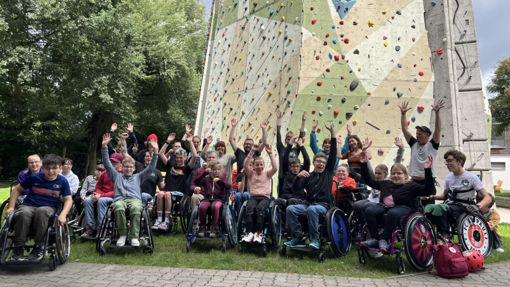 Gruppenfoto mit allen Teilnehmenden und Helfenden vor einer hohen Kletterwand in der DAV-Kletterhalle. Die Gruppe besteht aus Kindern, Jugendlichen und Erwachsenen – viele im Rollstuhl, alle lächeln in die Kamera.