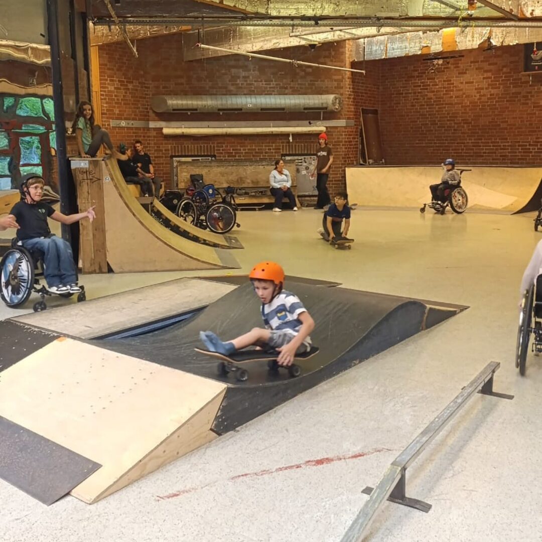Ein Junge sitzt auf einem Skateboard und fährt eine kleine Rampe im Skatepark des Ackerpoolco hinunter. Im Hintergrund sind weitere Kinder und Jugendliche im Rollstuhl, die sich in der Halle bewegen oder zuschauen.