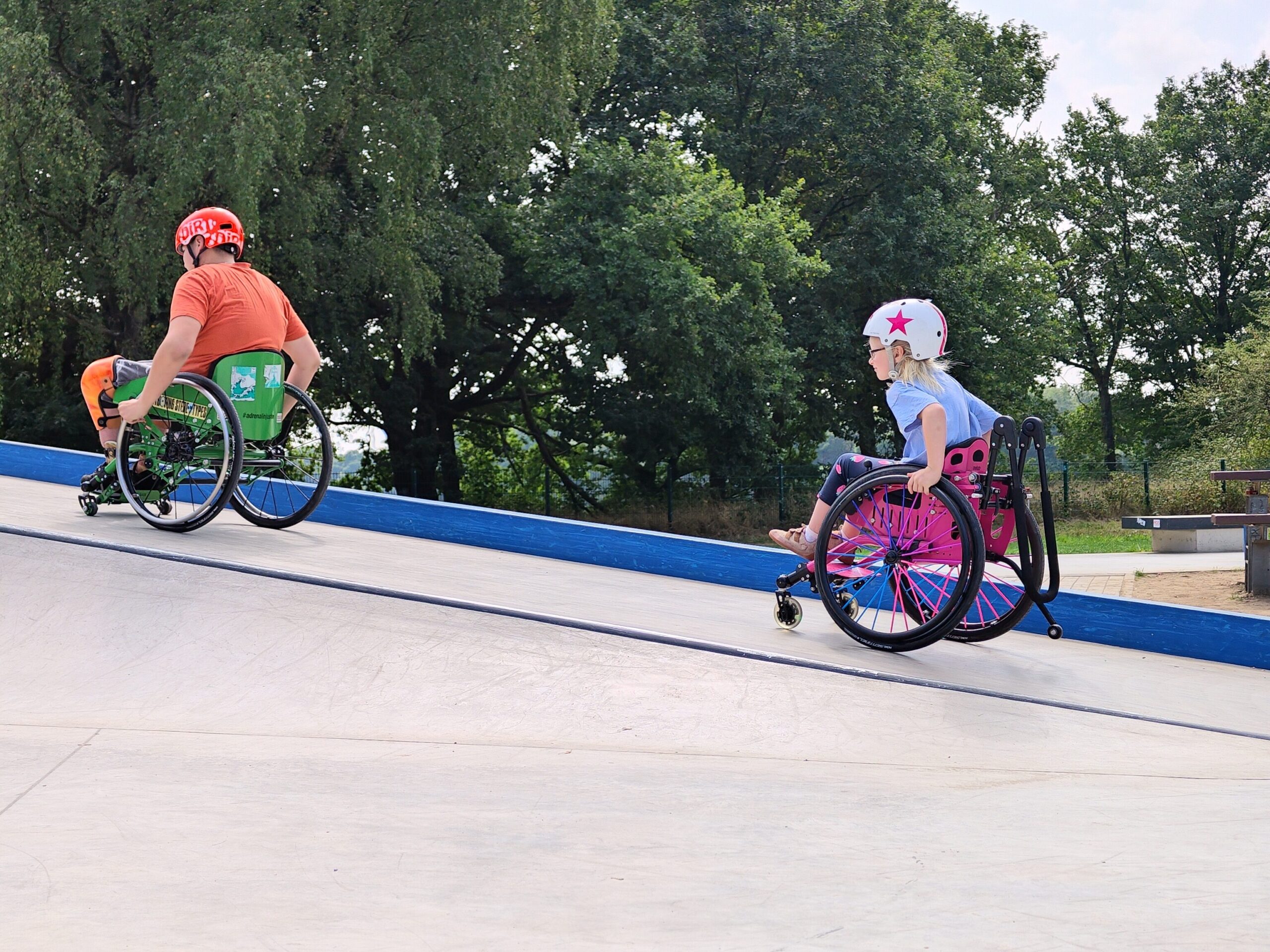 Zwei Kinder im Rollstuhl fahren nebeneinander eine schräge Ebene im Skatepark hoch. Links ein Junge mit orangem Shirt und Helm, rechts ein Mädchen mit weißem Helm mit rosa Stern und pinkem Rollstuhl. Im Hintergrund Bäume und Grünflächen.
