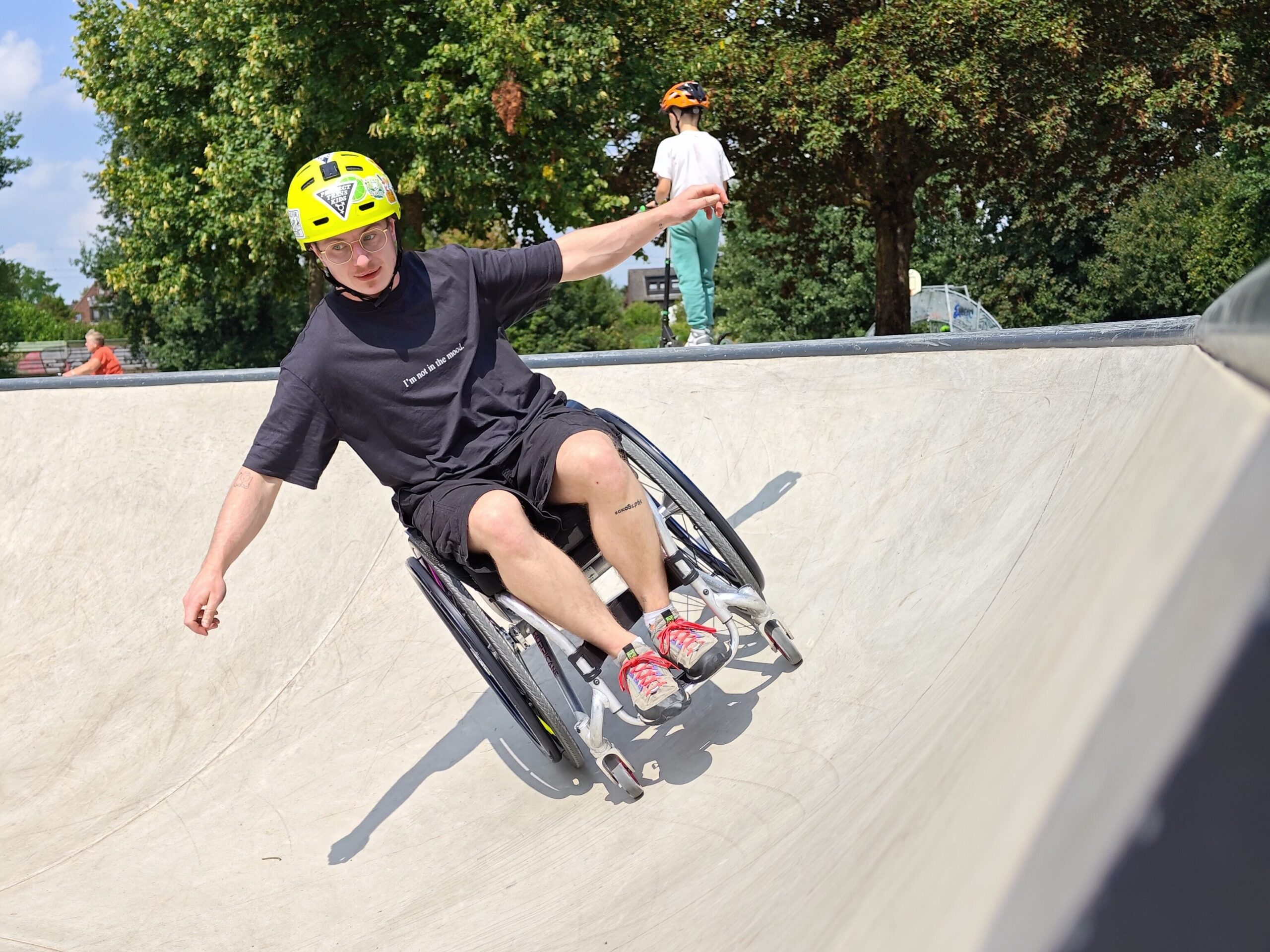 Ein junger Rollstuhlfahrender mit gelbem Helm fährt eine steile Betonrampe hinunter. Der rechte Arm ist ausgestreckt für Balance, der Gesichtsausdruck konzentriert. Im Hintergrund ist ein Kind mit Helm auf einem Skateboard zu sehen. Die Szene ist sonnig und aktiv.
