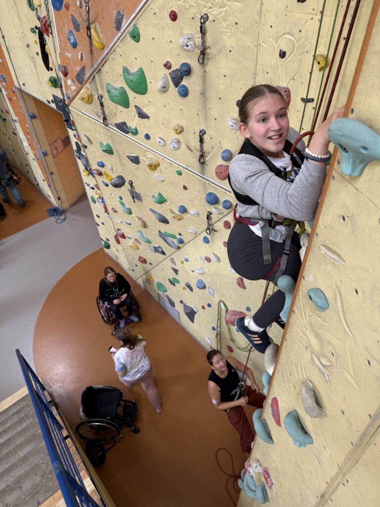 Eine jugendliche Teilnehmerin klettert konzentriert an der Kletterwand nach oben. Der Blick der Kamera ist von oben nach unten gerichtet. Unten im Bild stehen Helfende und eine weitere Rollstuhlfahrerin, die nach oben schauen und sichern.
