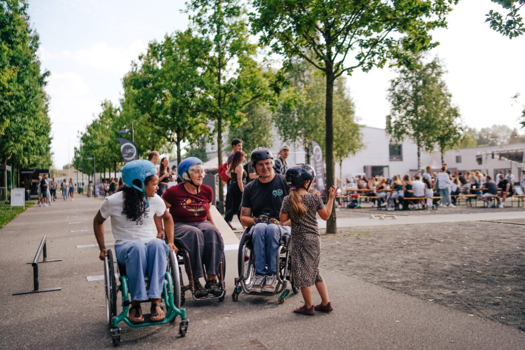 Eine Gruppe von vier Menschen sitzt in Rollstühlen auf einer Festivalstraße unter Bäumen – zwei Kinder, eine ältere Frau und ein Mann mit Helm. Alle tragen Schutzhelme und unterhalten sich lachend mit einem kleinen Mädchen im gepunkteten Kleid, das ebenfalls einen Helm trägt. Im Hintergrund sitzen Festivalgäste an Bänken und genießen das Programm.