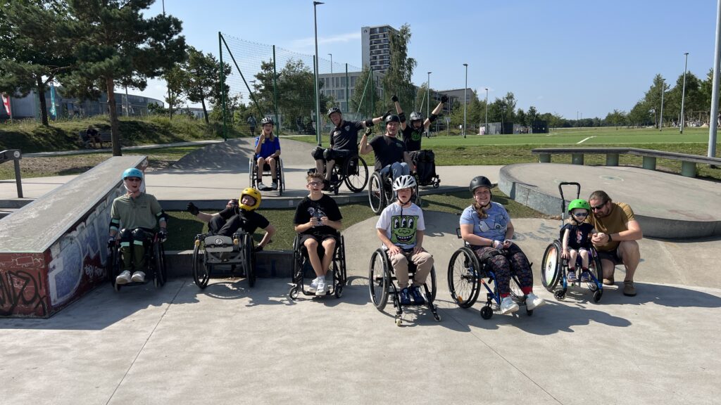 Gruppenfoto mehrerer Kinder und Jugendlicher im Rollstuhl im Skatepark. Sie sitzen in einer Reihe, lachen und posieren für die Kamera, manche reißen die Arme hoch.