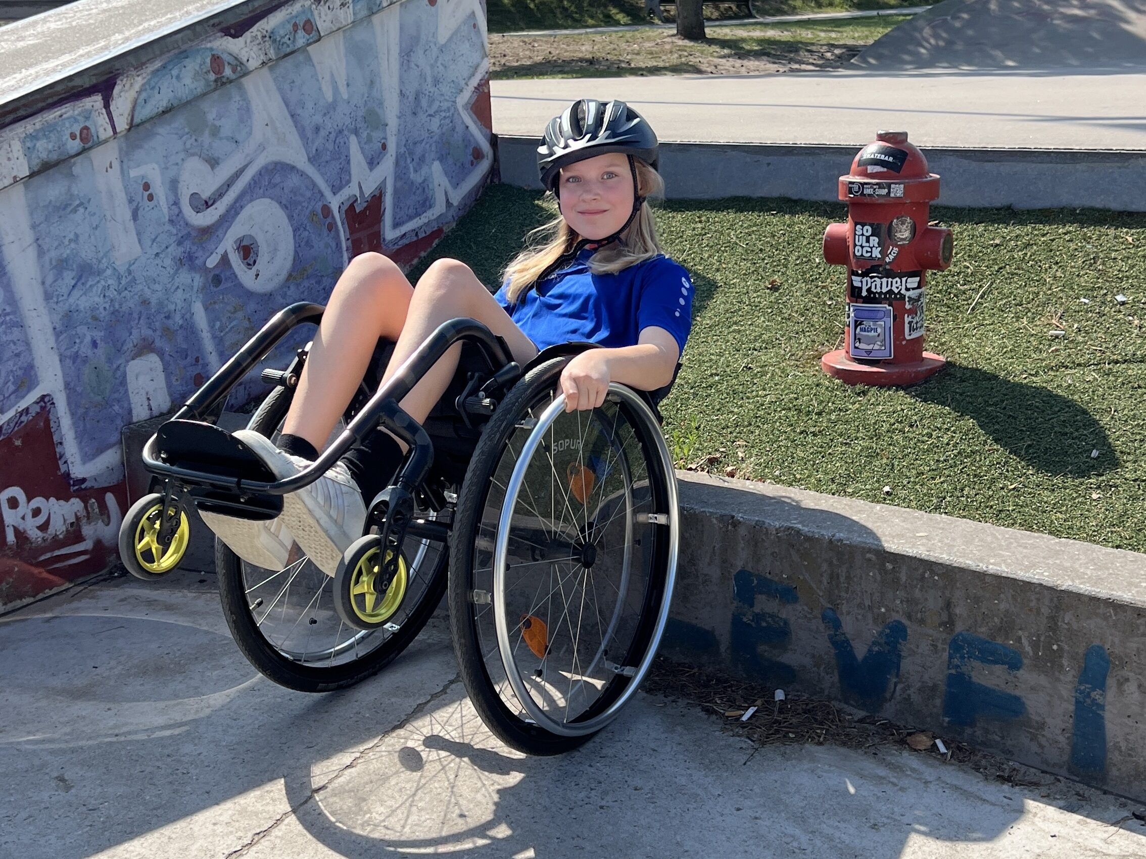 Eine Jugendliche sitzt lässig im Rollstuhl neben einer kleinen Rampe im Skatepark. Sie trägt Helm und lächelt in die Kamera, neben ihr steht ein roter Hydrant.