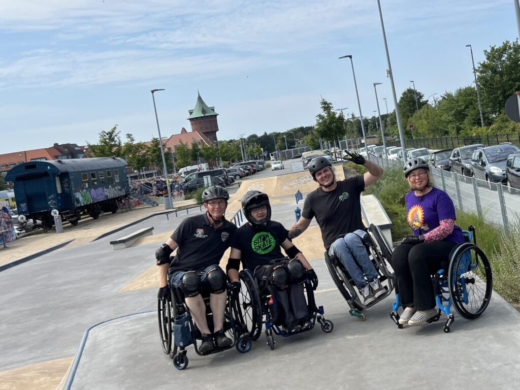 Vier Teilnehmende im Rollstuhl posieren nebeneinander auf einer Startfläche im Skatepark Cuxhaven. Alle tragen Helme und lachen in die Kamera, einer hebt die Hand zum Gruß.