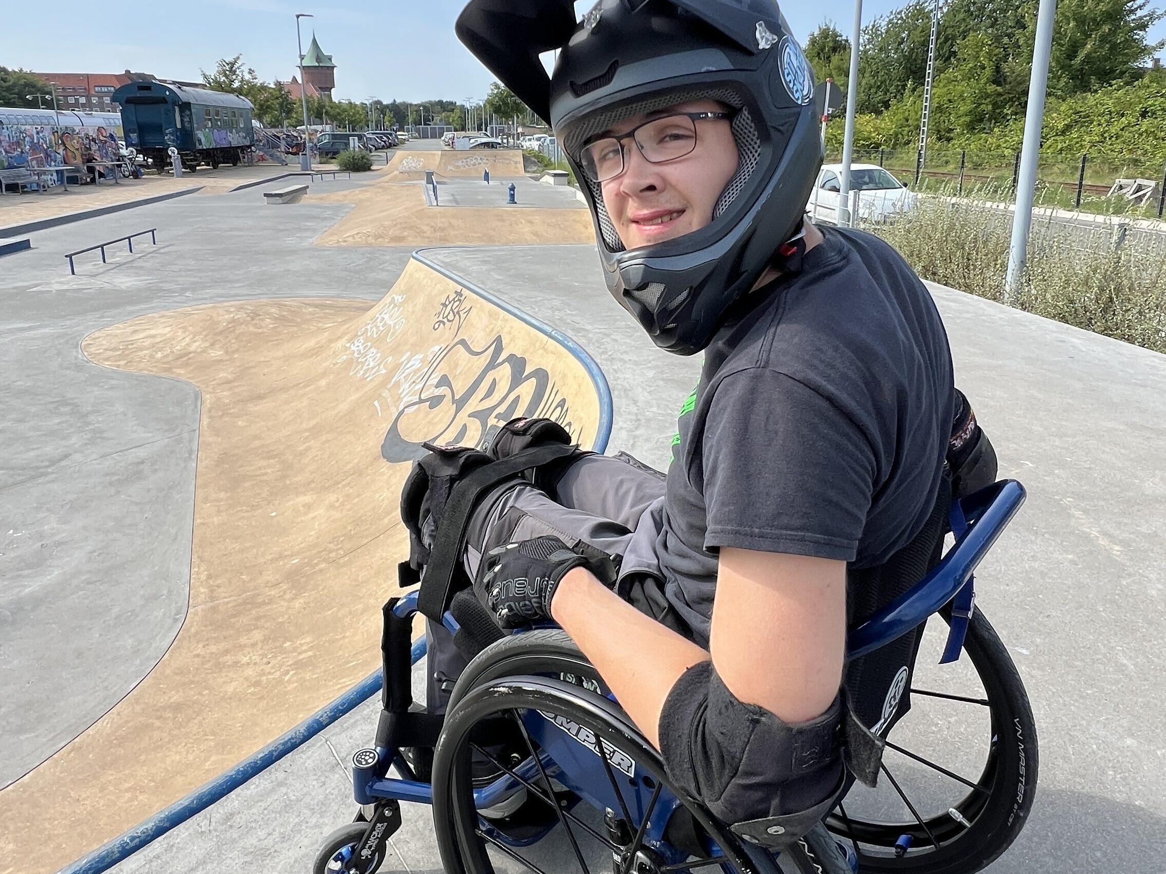 Ein Jugendlicher im Rollstuhl mit Helm und Schutzkleidung sitzt oben auf einer Rampe im Skatepark Cuxhaven und blickt in die Kamera. Im Hintergrund sind der Skatepark und ein markanter Turm sowie ein alter Zugwaggon zu sehen.