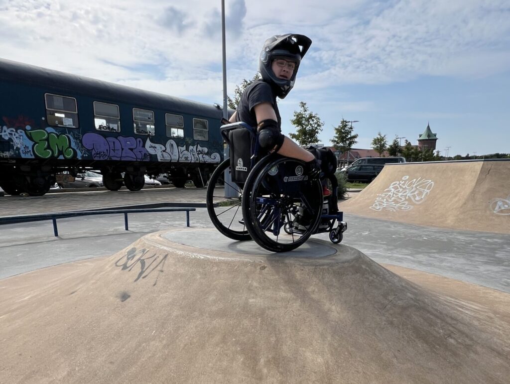 Ein Rollstuhlfahrender sitzt auf einem Hügel-Element im Skatepark Cuxhaven. Er trägt Helm und Schutzkleidung, im Hintergrund ist ein graffitibesprühter Zugwaggon zu sehen.