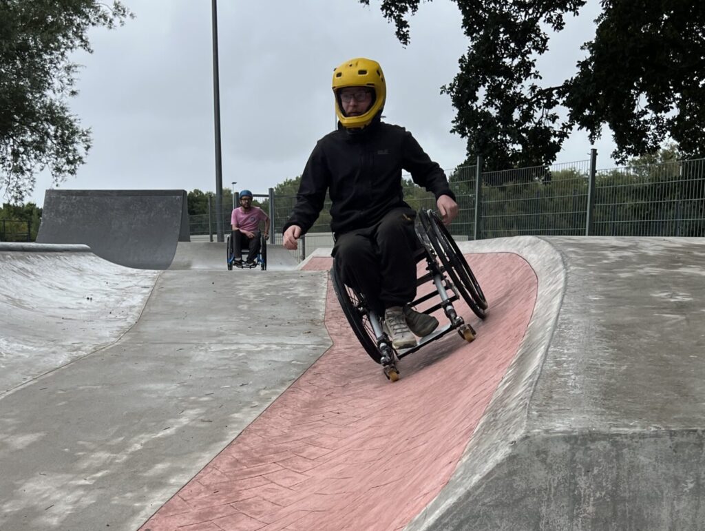 Ein Rollstuhlnutzender mit gelbem Helm fährt eine schräge, rote Fläche im Skatepark hinunter. Im Hintergrund sieht man eine weitere Person im Rollstuhl, die ebenfalls die Rampe befährt. Der Himmel ist grau, der Boden wirkt teilweise noch feucht.