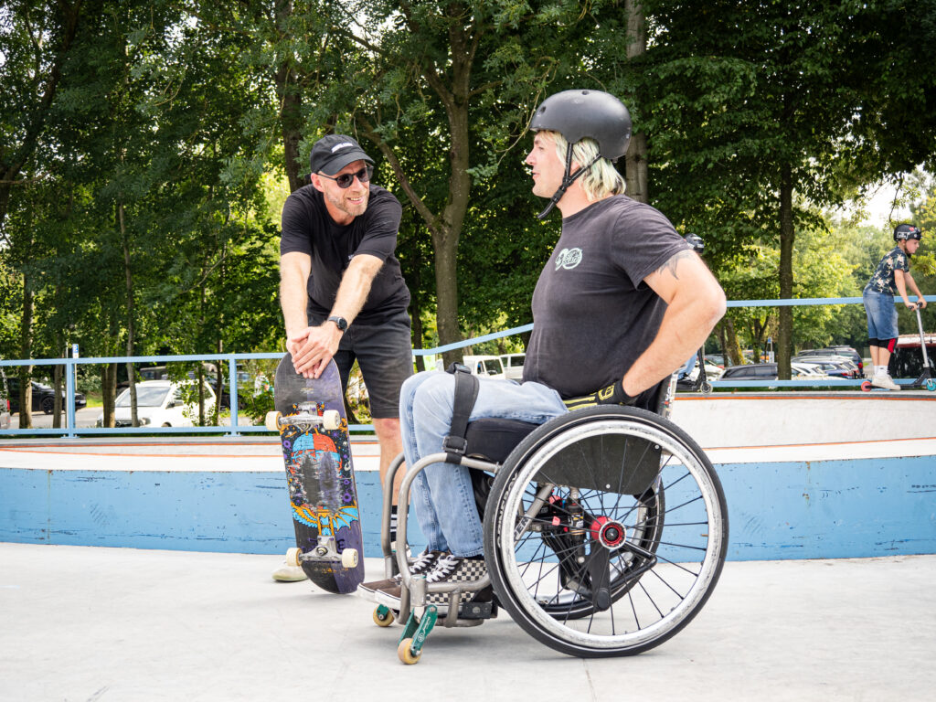 Ein Mann im schwarzen T-Shirt und Skatehelm sitzt im Rollstuhl im Skatepark und unterhält sich mit einem Skateboarder, der sein Board vor sich hält. Beide wirken konzentriert und freundlich, im Hintergrund sind weitere Menschen auf Scootern und Skateboards zu sehen.