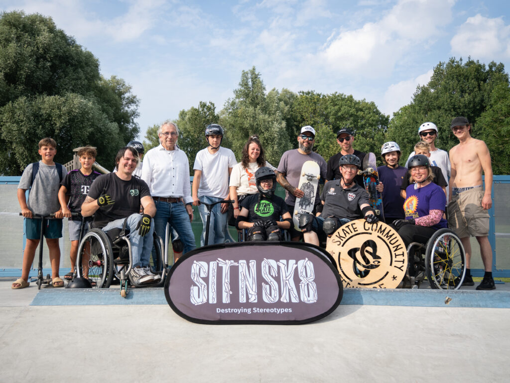 Gruppenfoto im Skatepark: Erwachsene, Kinder und Jugendliche mit und ohne Rollstuhl posieren gemeinsam für ein Bild. Vorne steht ein Banner mit dem SIT’N’SKATE-Logo und dem Schriftzug „Destroying Stereotypes“. Alle lächeln und wirken stolz.