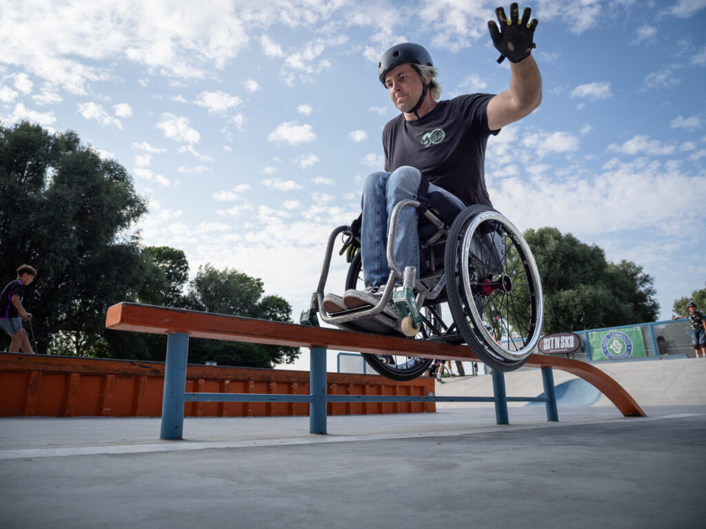 Ein Rollstuhlfahrender fährt im Skatepark über ein Rail und hebt dabei die Hand. Er trägt Helm und Handschuhe, die Szene wirkt dynamisch, mit blauem Himmel und einigen Zuschauenden im Hintergrund.
