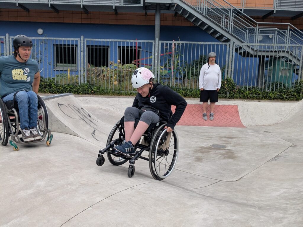 Zwei junge Menschen im Rollstuhl fahren im Skatepark. Im Vordergrund eine Person mit weißem Helm, die sich konzentriert in die Kurve legt. Eine weitere Person mit schwarzem Helm beobachtet. Im Hintergrund steht eine Begleitperson vor einem Schulgebäude mit orangefarbener Fassade.