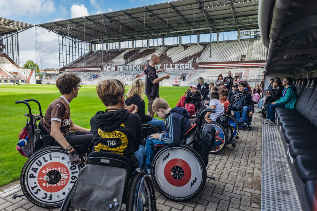 Mehrere Kinder und Jugendliche im Rollstuhl sitzen am Spielfeldrand im Millerntor-Stadion. Eine Person steht vor ihnen und erklärt etwas, im Hintergrund die leeren Tribünen mit dem Schriftzug „Kein Mensch ist illegal“.