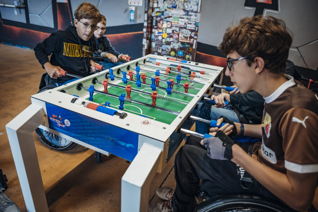 Vier Jugendliche im Rollstuhl spielen an einem Kickertisch. Alle sind konzentriert im Spiel. Im Hintergrund bunte Sticker und Poster.