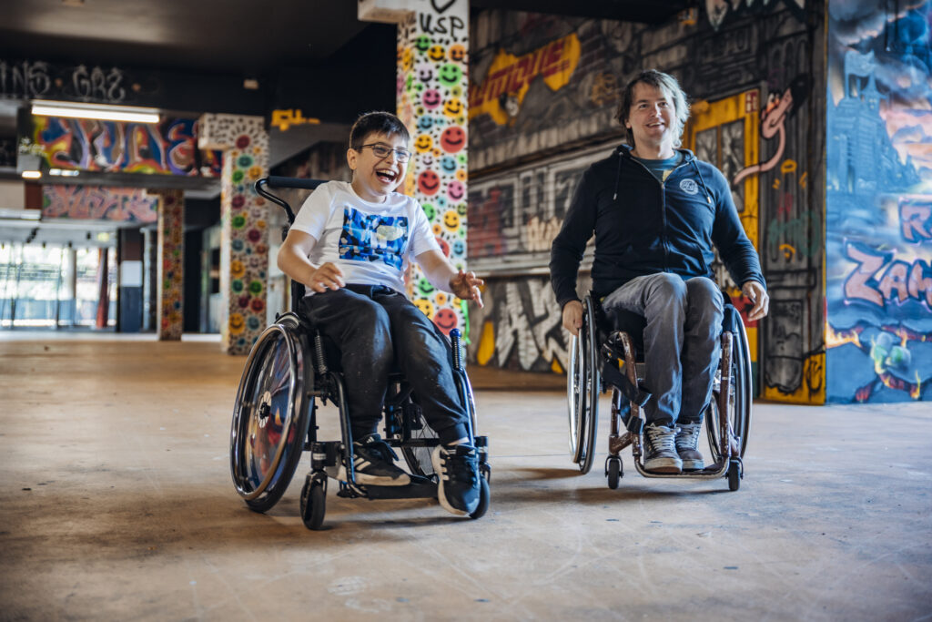 Ein Kind und ein Erwachsener rollen gemeinsam lachend im Rollstuhl durch einen bunt bemalten, graffiti-verzierten Gang im Stadion.
