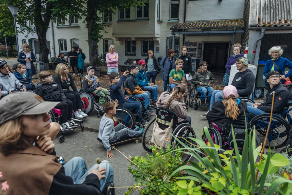 Eine große Gruppe von Kindern, Jugendlichen und Helfenden – viele davon im Rollstuhl – versammelt sich im Hof des Goldbekhauses. Alle hören aufmerksam zu, einige lächeln. Im Hintergrund stehen weitere Erwachsene.
