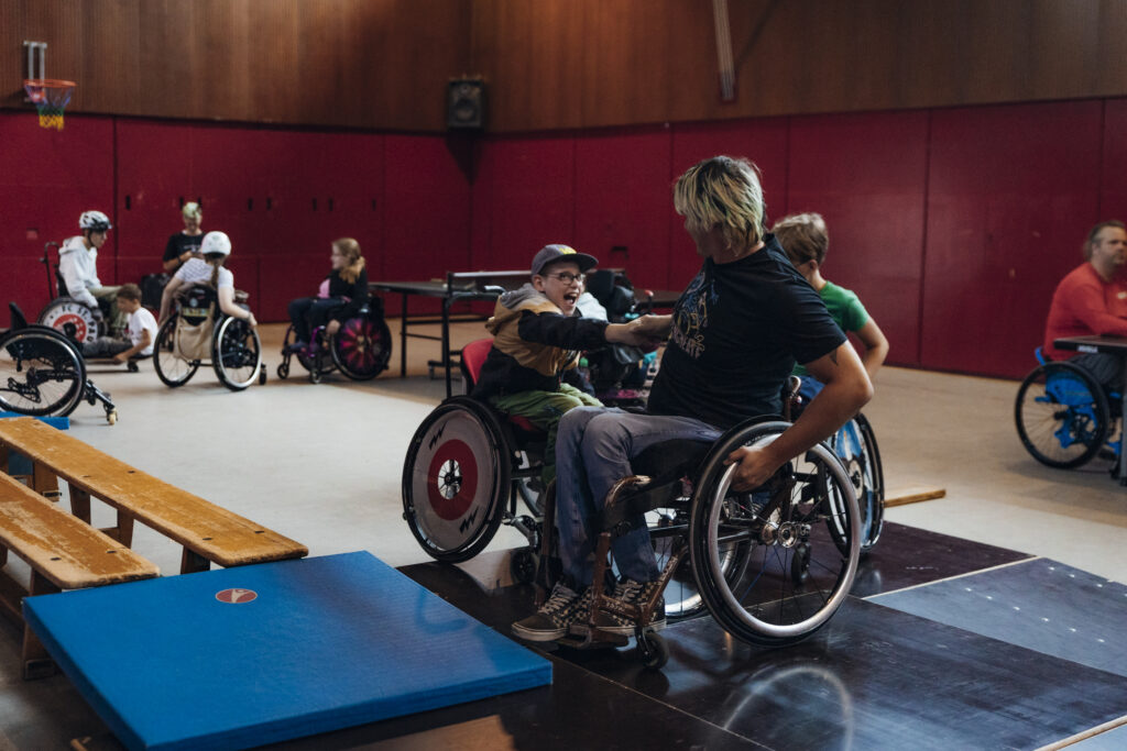 Zwei Kinder im Rollstuhl in einer Sporthalle lachen sich an und haben offensichtlich Spaß. Im Hintergrund weitere Kinder beim Rollstuhltraining oder Tischtennisspielen. Vorne liegen Sportmatten und Bänke.
