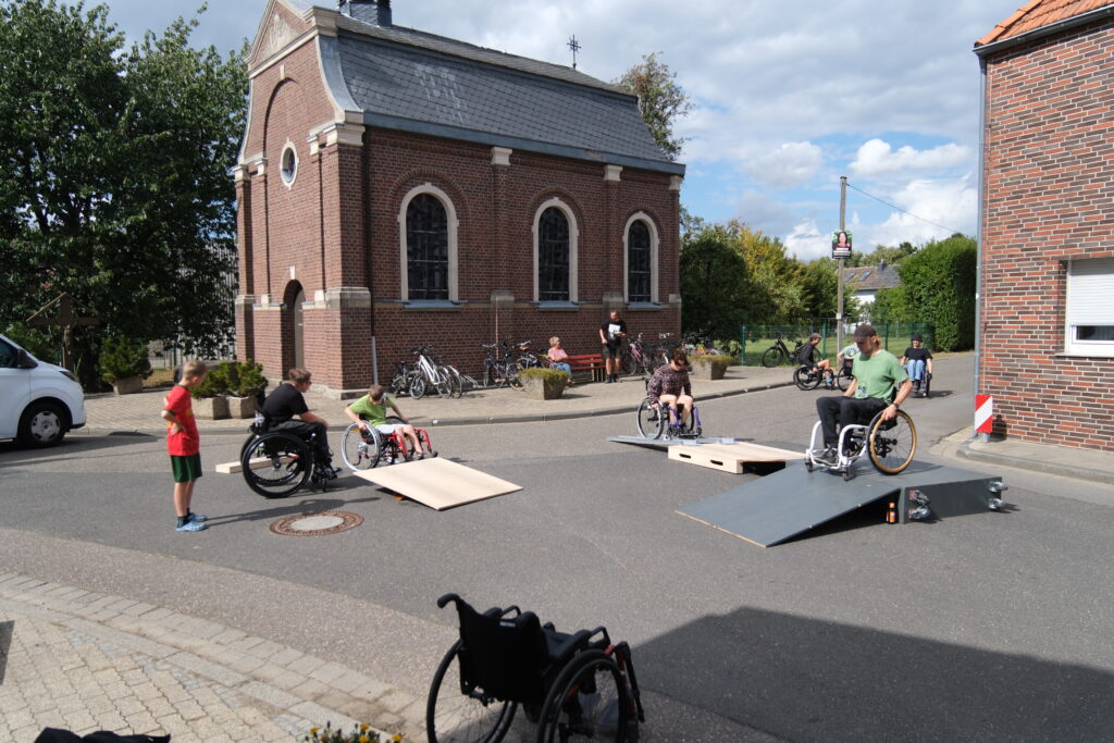 Szene des Rolliparcours vor einer kleinen Kirche. Kinder und Erwachsene im Rollstuhl nutzen verschiedene Rampen, während einige Zuschauende am Rand stehen oder auf einer Bank sitzen. Die Stimmung ist entspannt und aktiv zugleich.