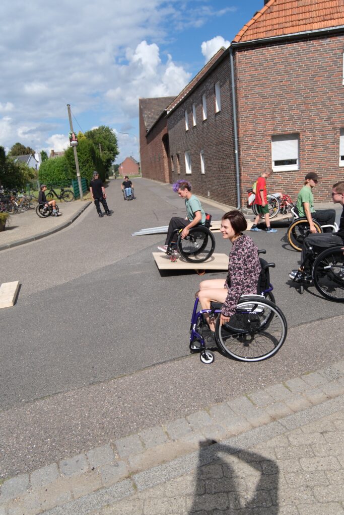Mehrere junge Menschen im Rollstuhl fahren über einen Rolliparcours auf einer ruhigen Dorfstraße. Die Teilnehmenden wirken konzentriert, einige lächeln. Die Strecke ist gesäumt von Backsteinhäusern und Bäumen, der Himmel ist blau mit einigen Wolken.