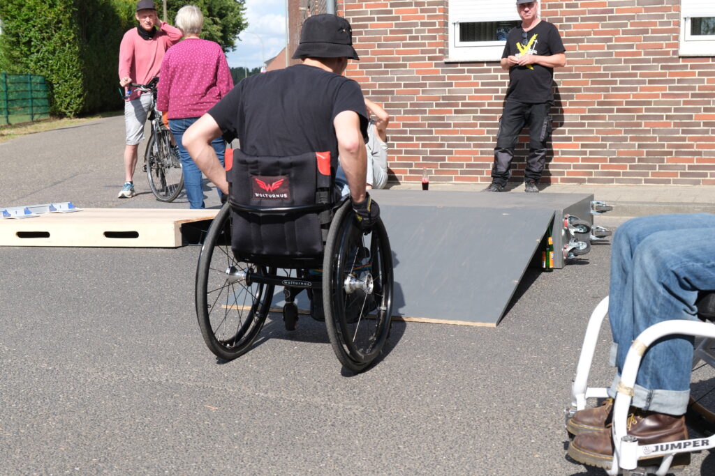 Ein Rollstuhlfahrender nähert sich einer mobilen Rampe auf der Straße. Die Rampe ist Teil eines Rolliparcours. Im Hintergrund unterhalten sich zwei Menschen mit einem Fahrrad vor einer Backsteinwand, ein weiterer steht daneben mit einem Getränk in der Hand.